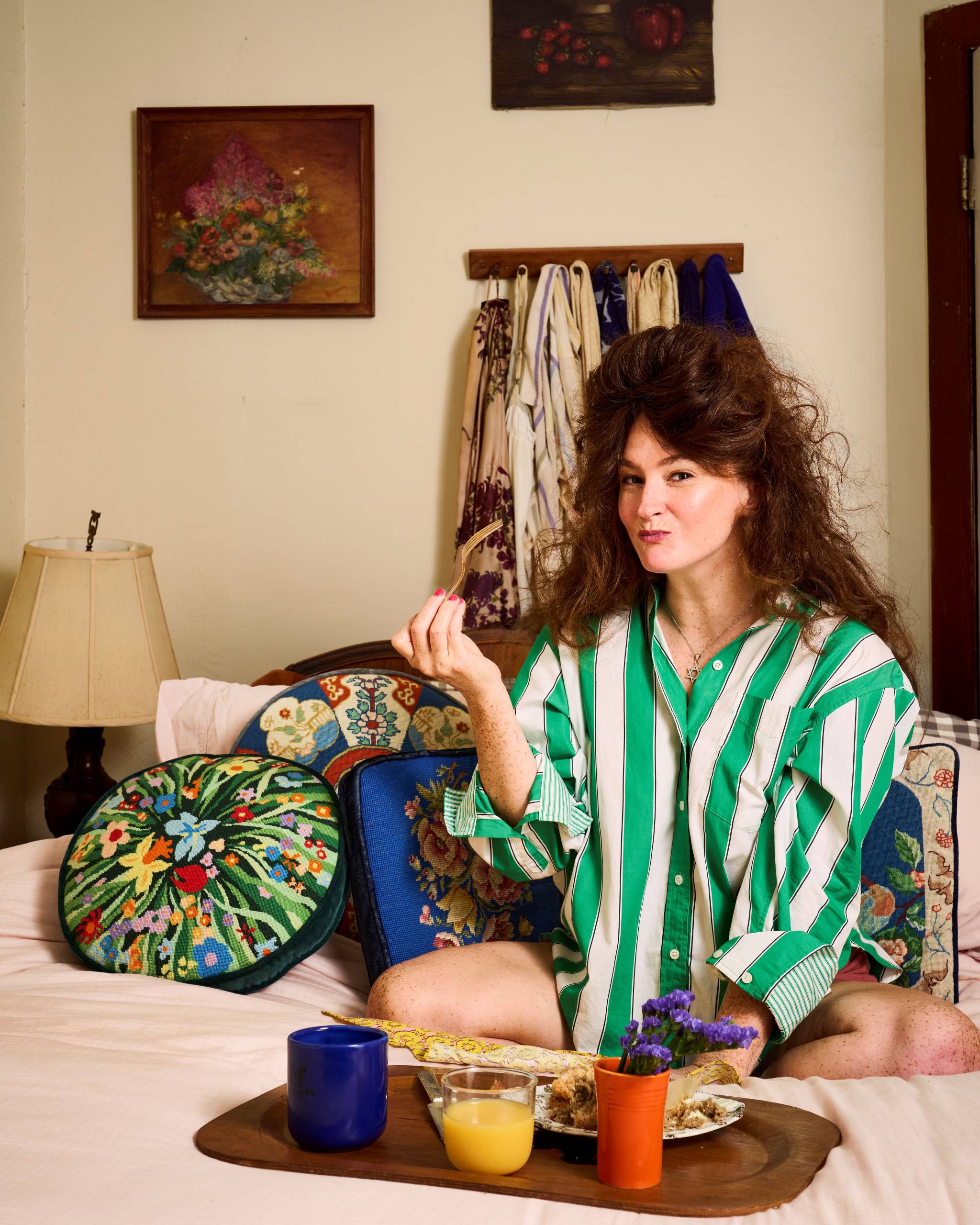 Woman with green striped shirt sits on bed in cozy room with flowers, drinks, and robes.