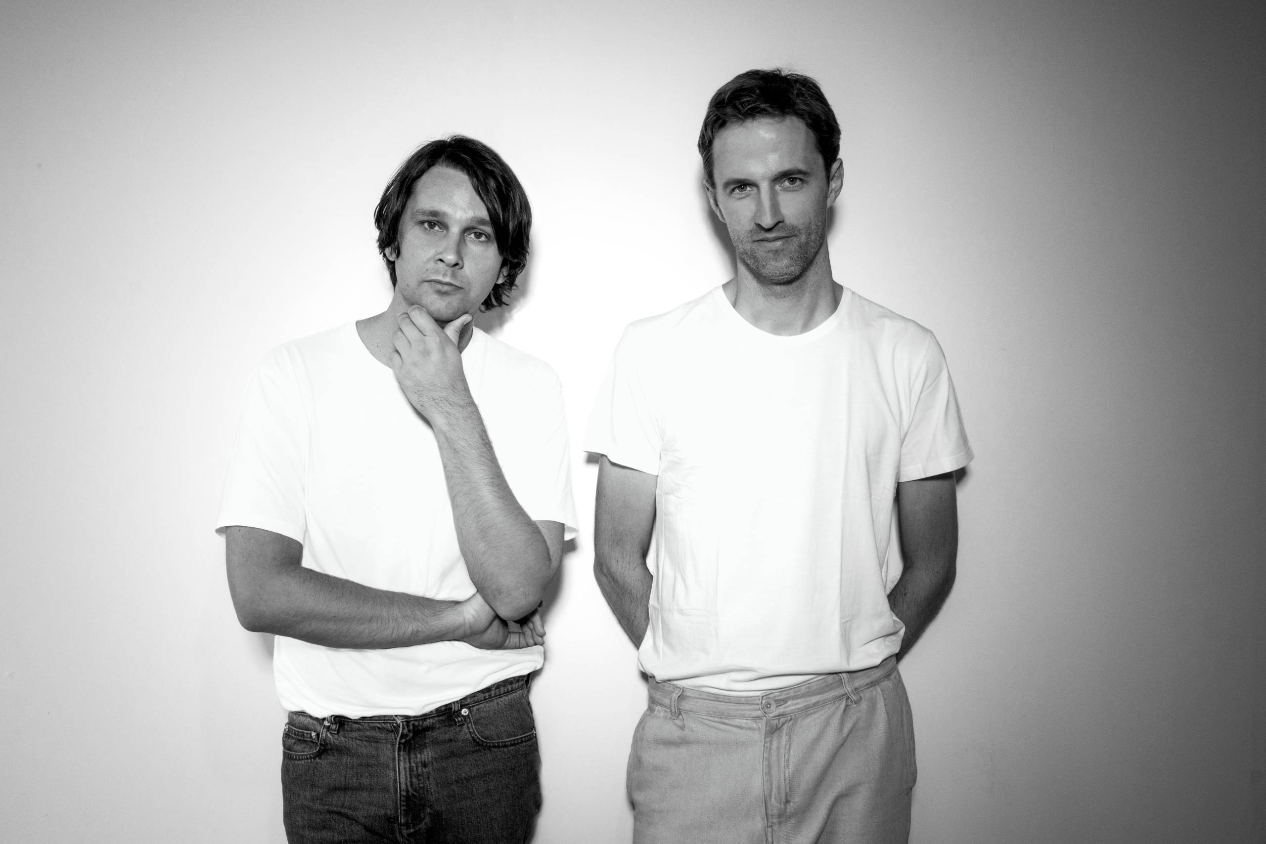Two men in white shirts stand against a plain background, one touching his chin.