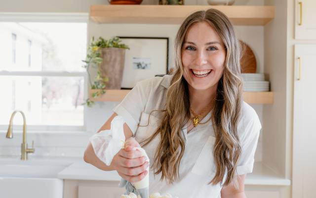 Woman icing a tall white cake in a bright kitchen, frosting with a piping bag.