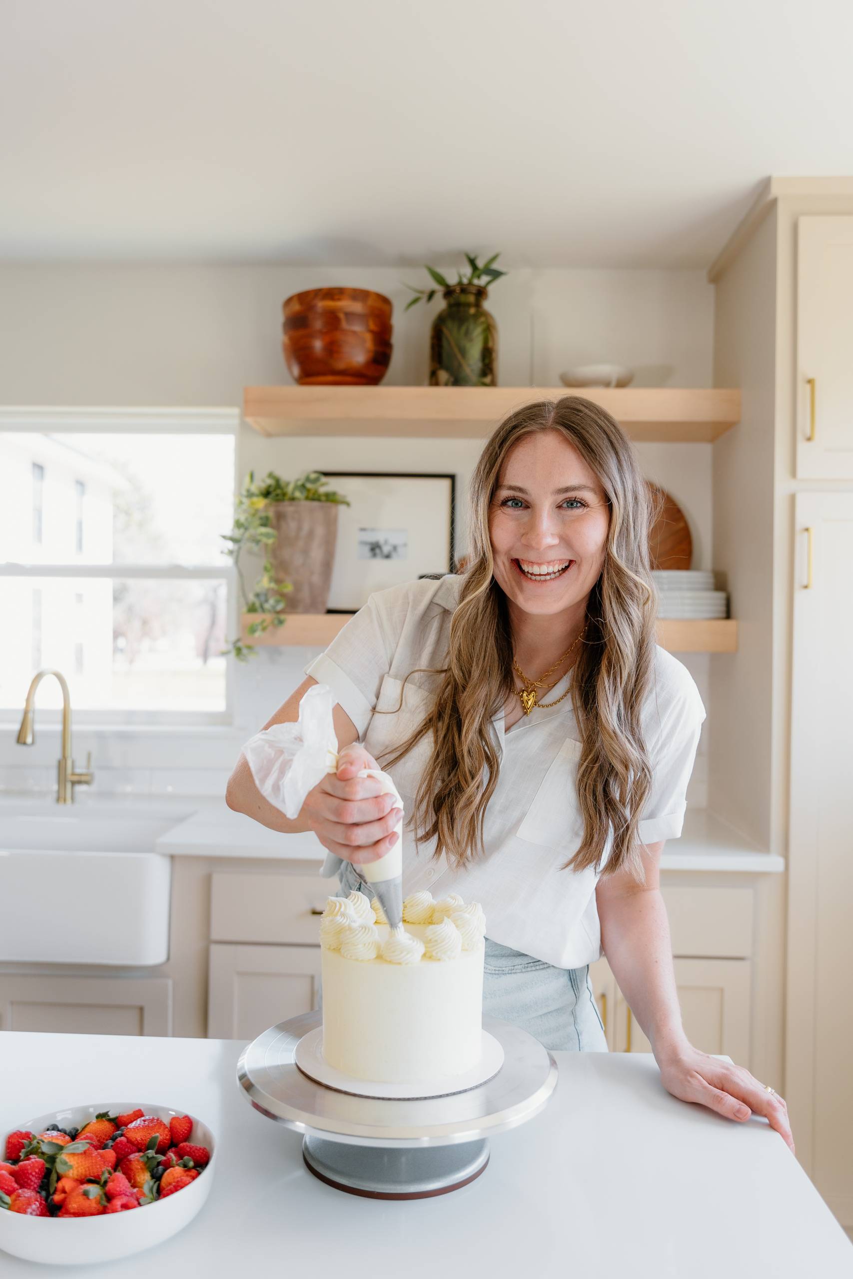Woman icing a tall white cake in a bright kitchen, frosting with a piping bag.