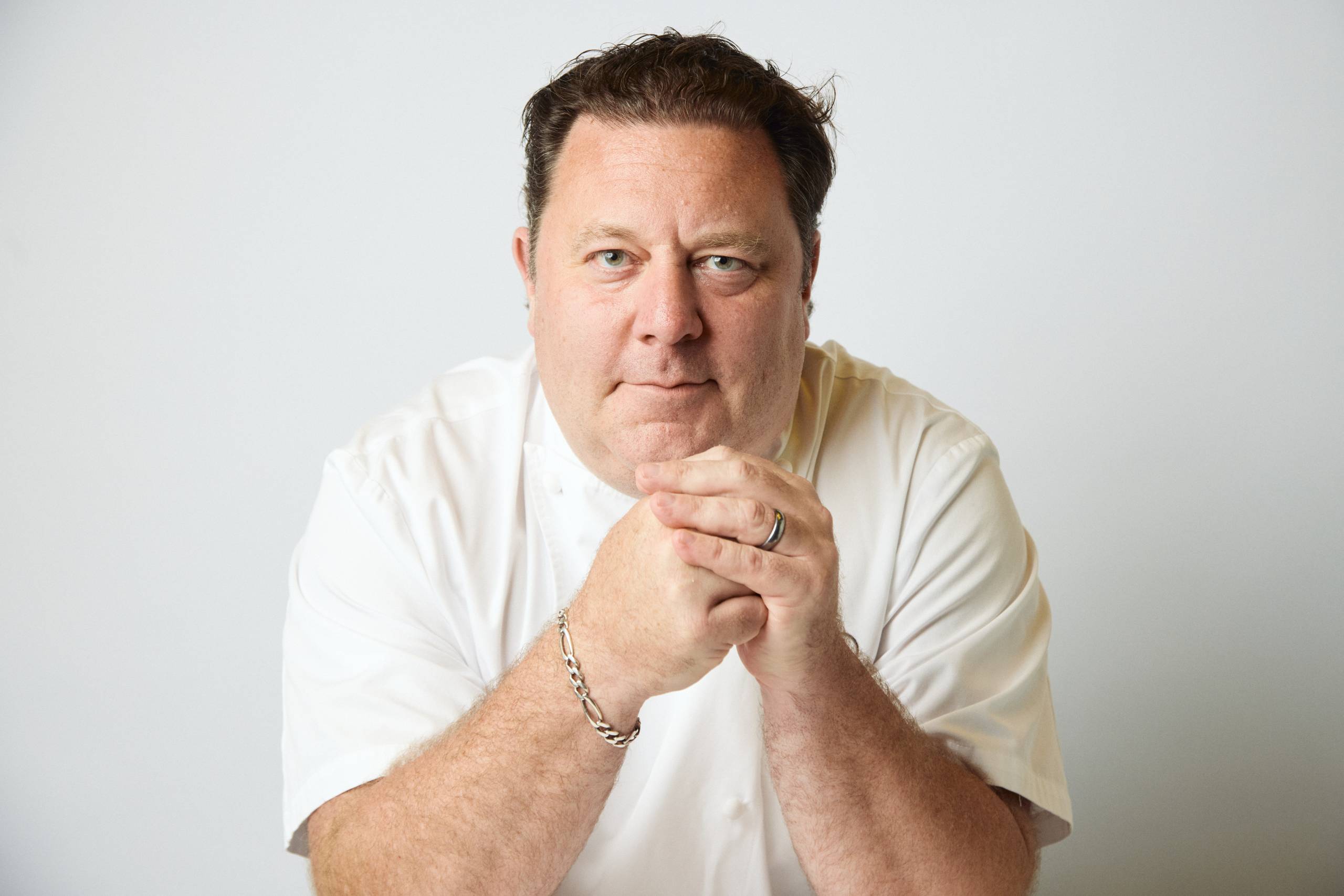 Middle-aged man with short dark hair in white shirt, hands clasped, neutral expression.