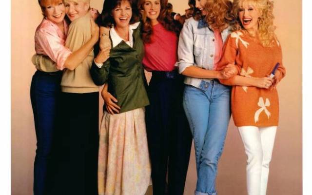 Six women standing close, smiling, arms around each other in colorful outfits.