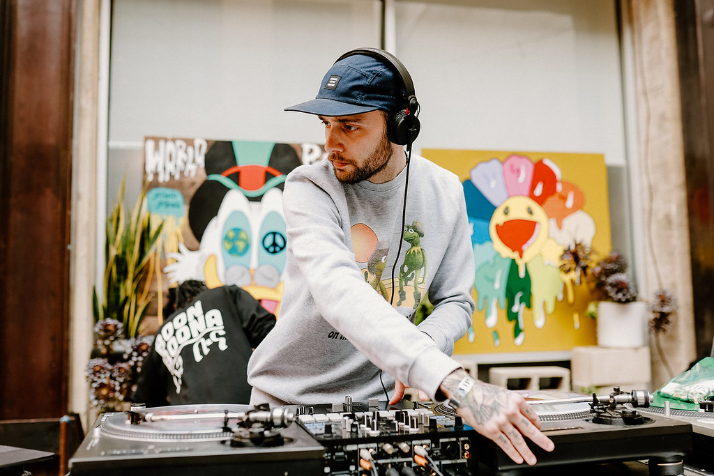 DJ wearing cap and headphones operating turntables indoors with colorful wall art behind.