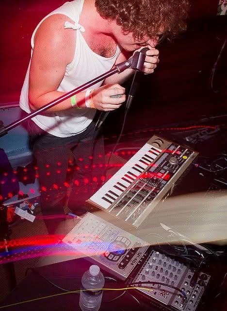 Male musician in white sleeveless shirt singing into microphone with synth and gear on stage.