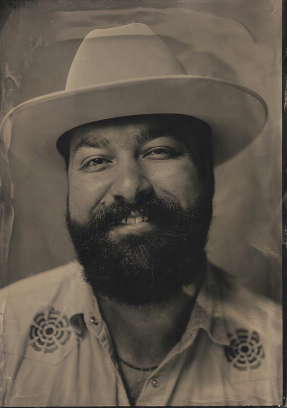 Man with full beard and mustache wearing a wide-brimmed hat, light embroidered shirt.