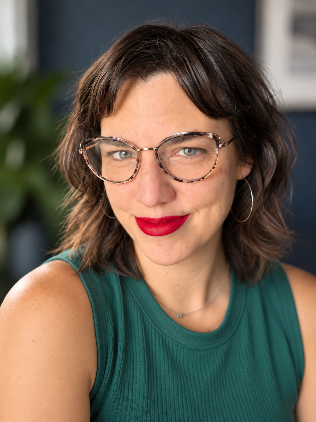 Woman with short brown hair, tortoiseshell glasses, teal sleeveless top, red lipstick.