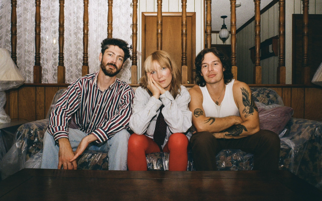 Three friends sit on a plastic-covered couch in a retro room, posing for the camera.
