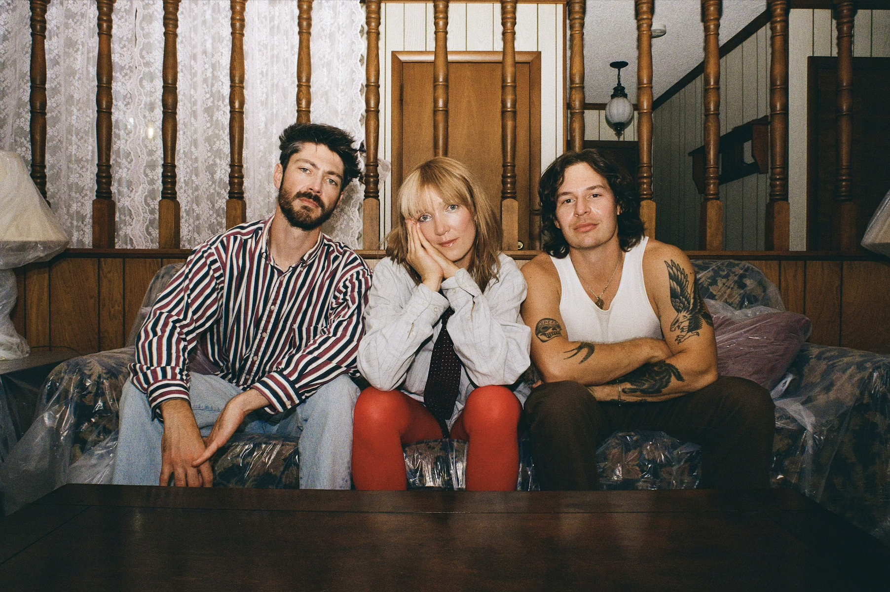 Three friends sit on a plastic-covered couch in a retro room, posing for the camera.