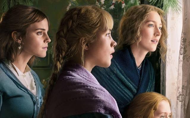 Four young women in sweaters stand by a window with Christmas greenery and decorations.